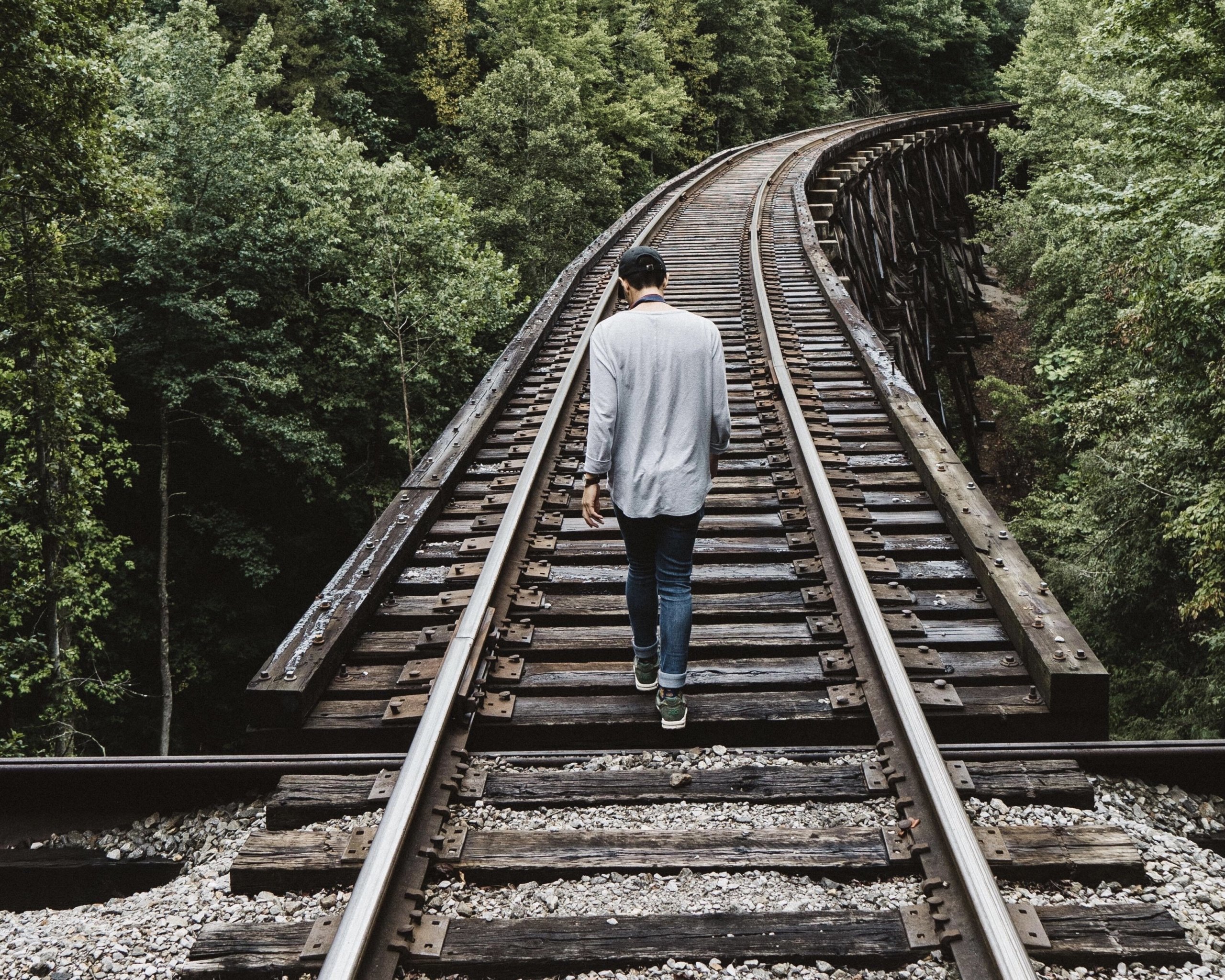 View from behind of a man wearing a white vest and jeans, walking down the middle of railroad tracks that wind through a forest