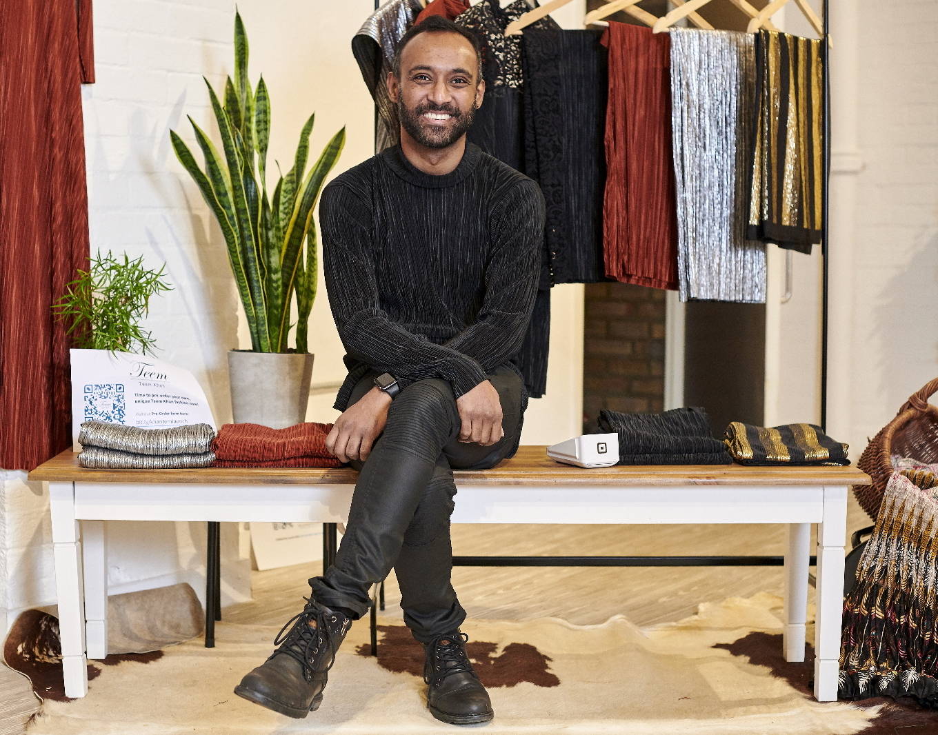 Teem Khan the fashion designer a dressed in black and smiling and sitting on a wooden bench amid clothing that the has designed with green plants in the background