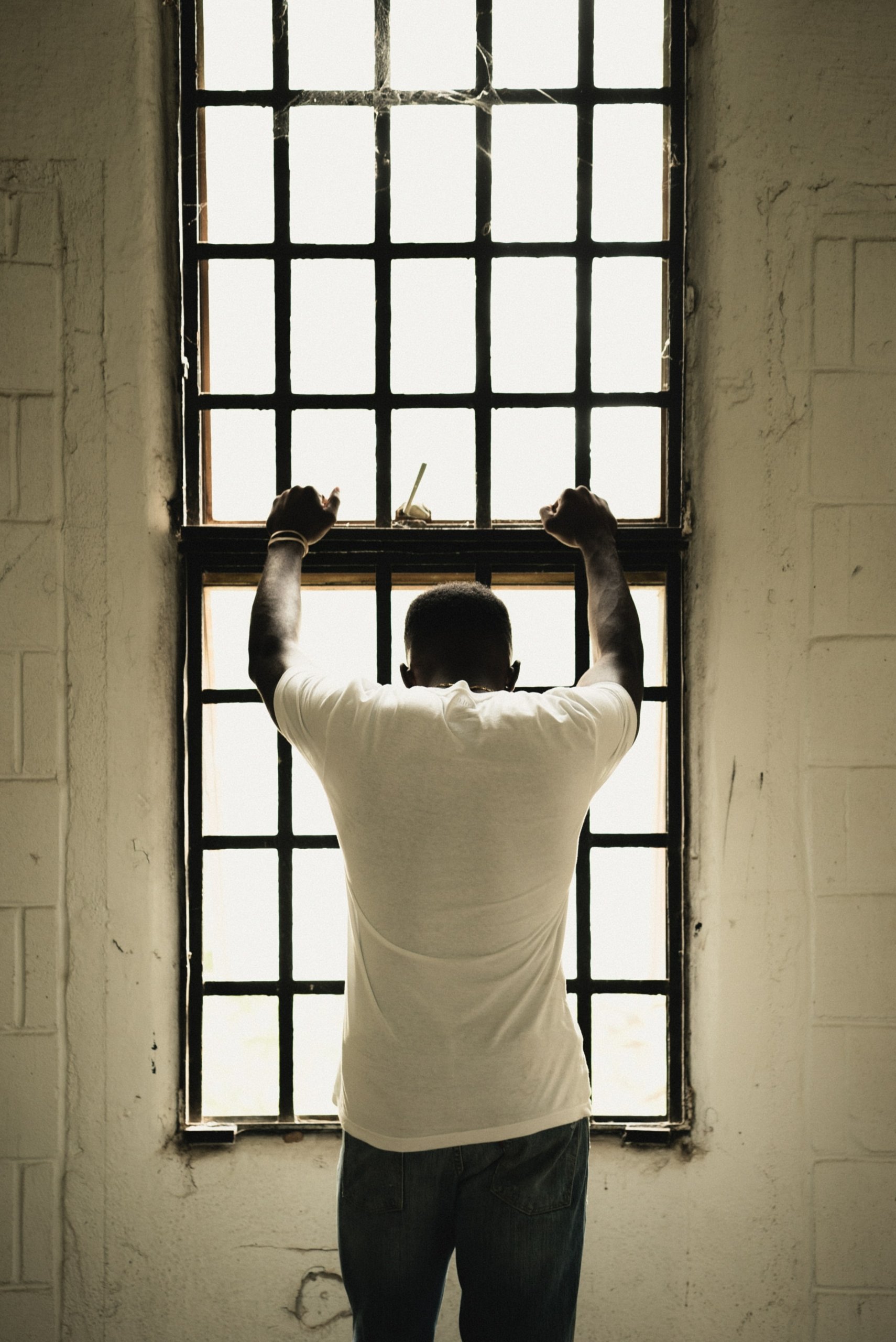 A man in a white tee shirt standing holding the bars of a window that he his facing 