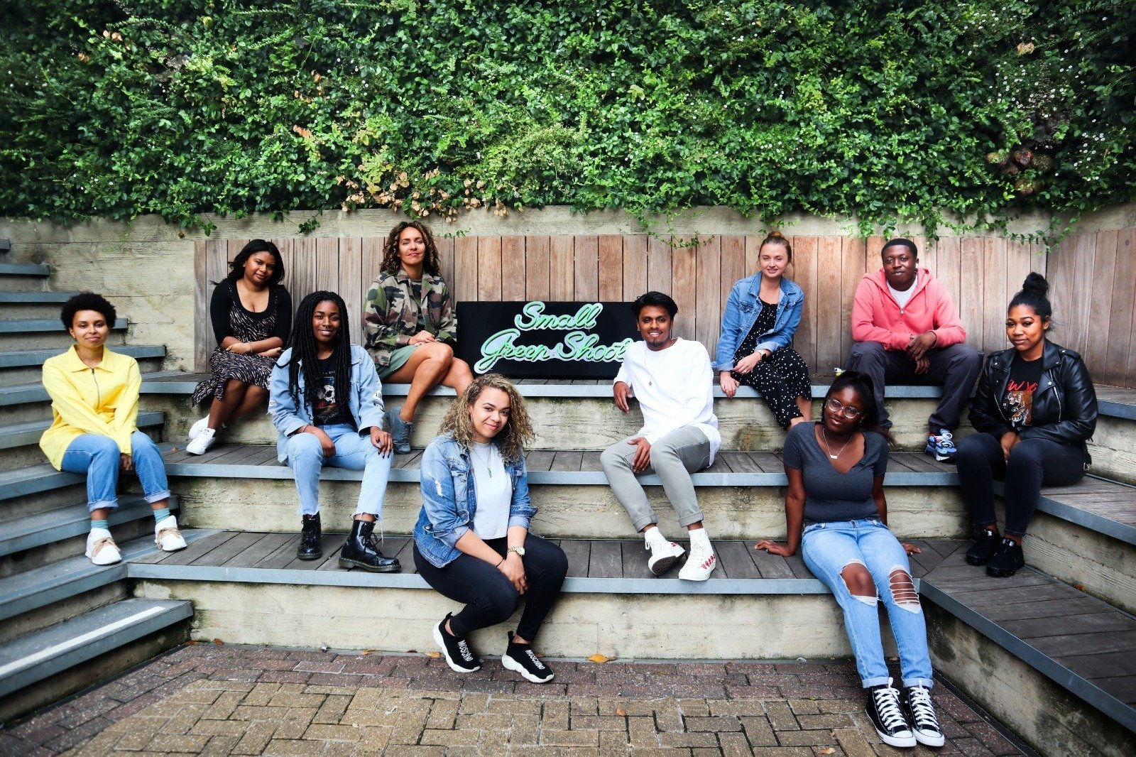 A group of 10 young people involved with place based giving at Camden Giving sitting outside on large, wood steps in an urban setting against a backdrop of a wood fence and a wall of green bushes with a signs that reads "small green shoots"