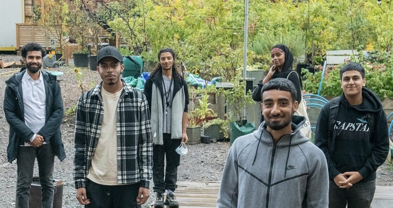 A group of 7 young people involved with place based giving at Camden Giving standing outside in an urban setting against a backdrop of plants, trees and bushes.