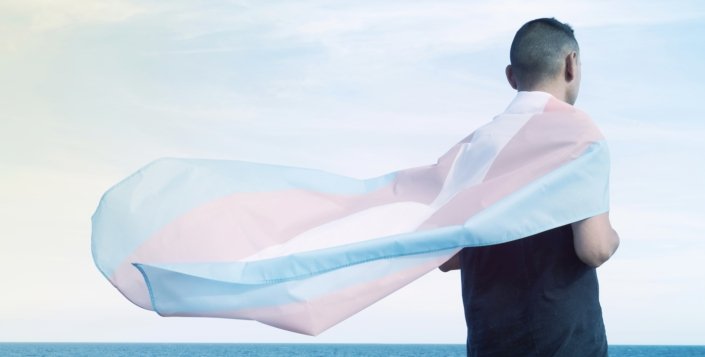 TransActual closeup of a person seen from behind wrapped in a Transgender Flag worn as a cape