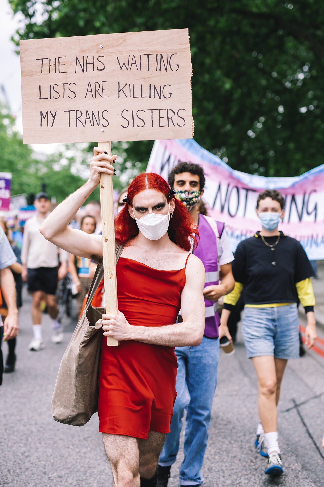 TransActual a person in a short red dress, wearing a facemask and carry a sign that reads "The NHS waiting lists are killing my trans sisters" as part of the London Trans Pride parade