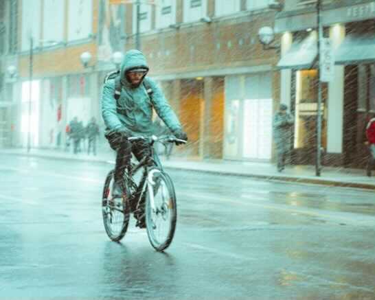 Bike Brigade featured image of a bearded cyclist wearing a hooded jacket and back pack speeds through the pouring rain as pedestrians walk down a sidewalk of an urban street