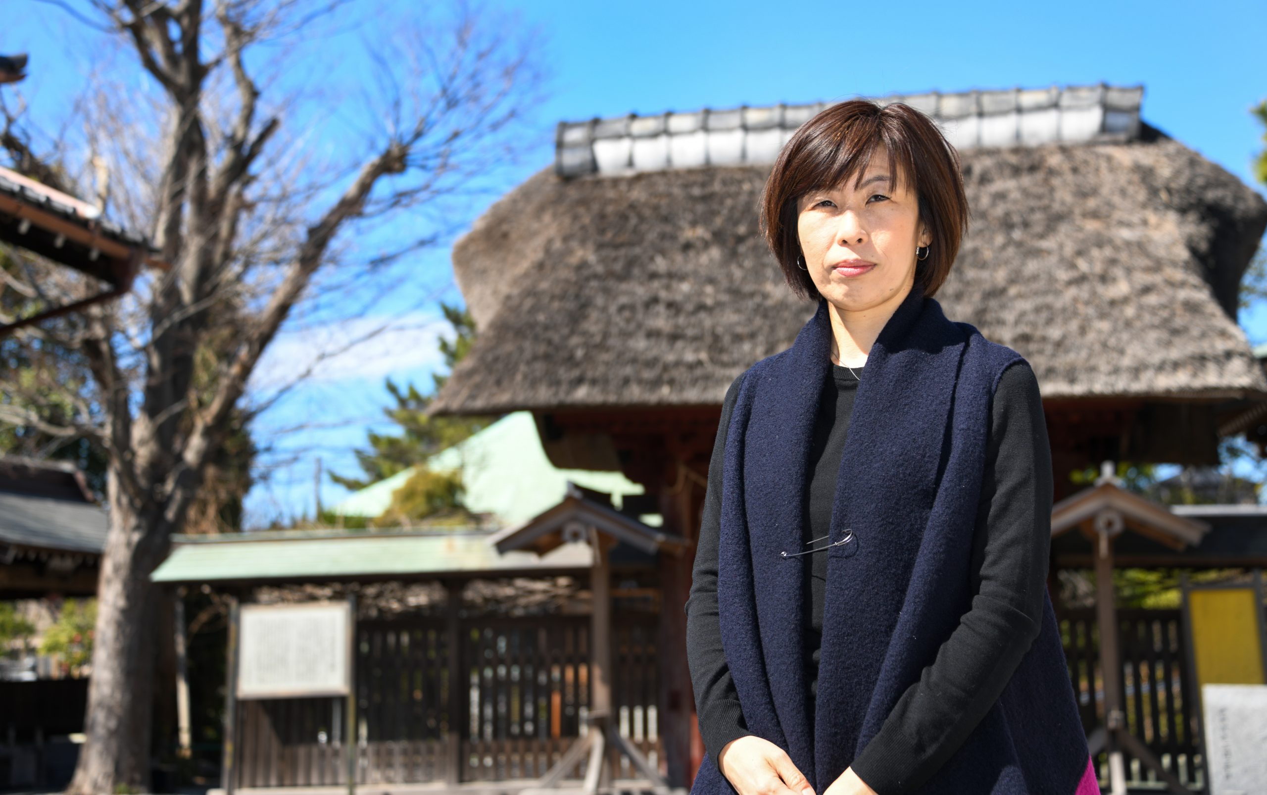 Anti Coal Power activist Kimiko Hirata standing outside on a sunny day in front a a building with a thatched roof and a tree whose leaves have fallen