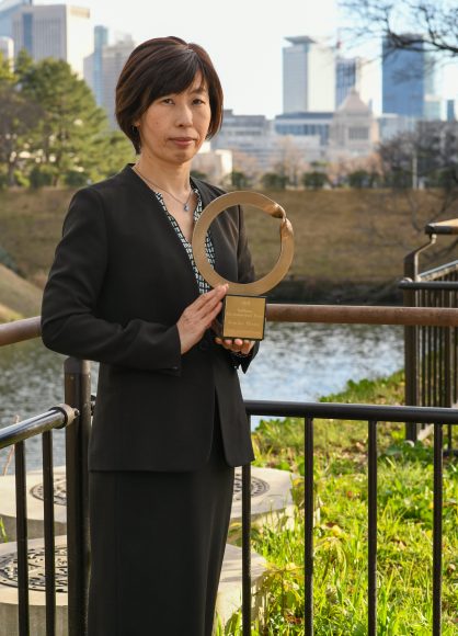 Goldman Environmental Prize winner Kimiko Hirata standing outside in front of a fence and against an urban backdrop of tall buildings as she holds the 2021 Ouroboros trophy