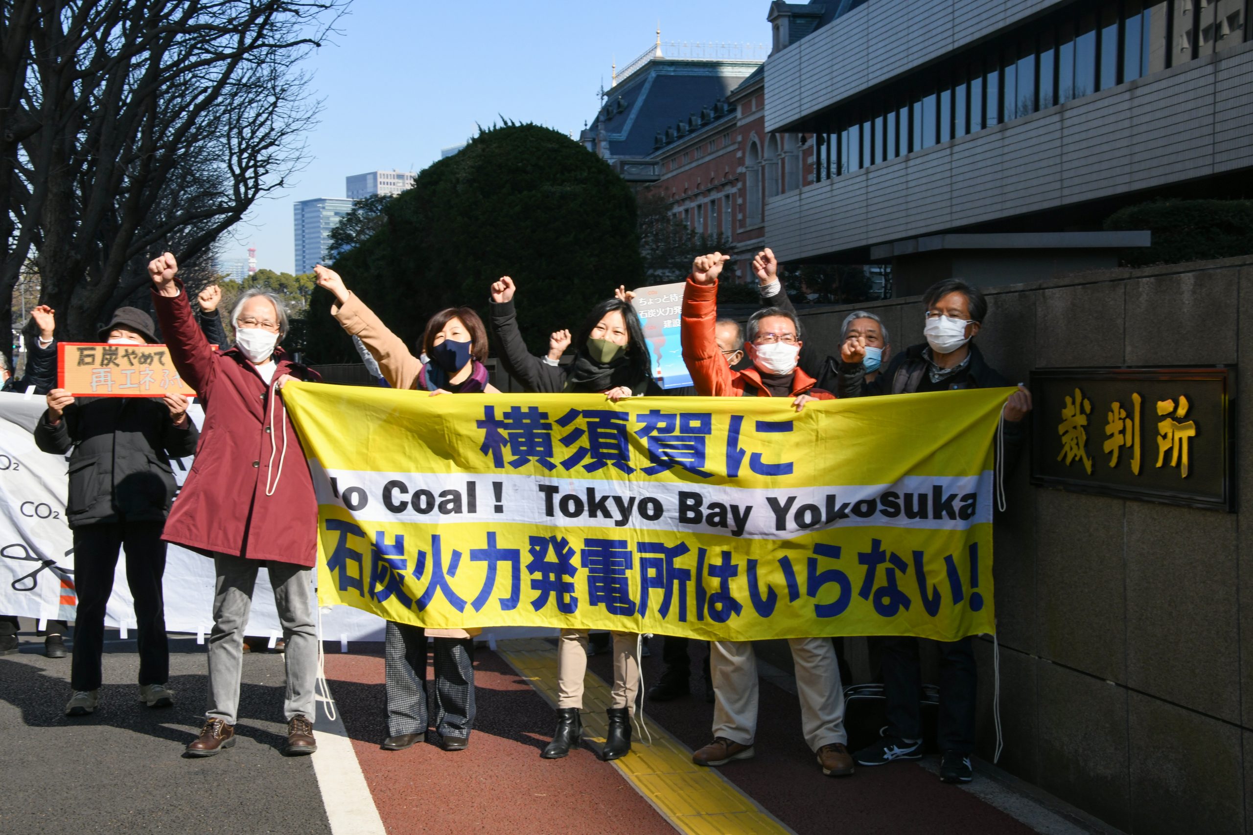 A group of people protesting against coal power wearing face-masks march on a street and raising their right fists in the air hold various signs including a yellow banner in Japanese and English reading (in English) No Coal ! Tokyo Bay Yokosuka Protest