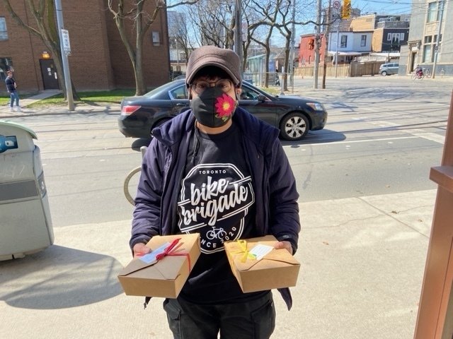 A masked person wearing a hat and a Bike Brigade tee shirt standing in the street and holding a delivery box in the hand of each outstretched arm
