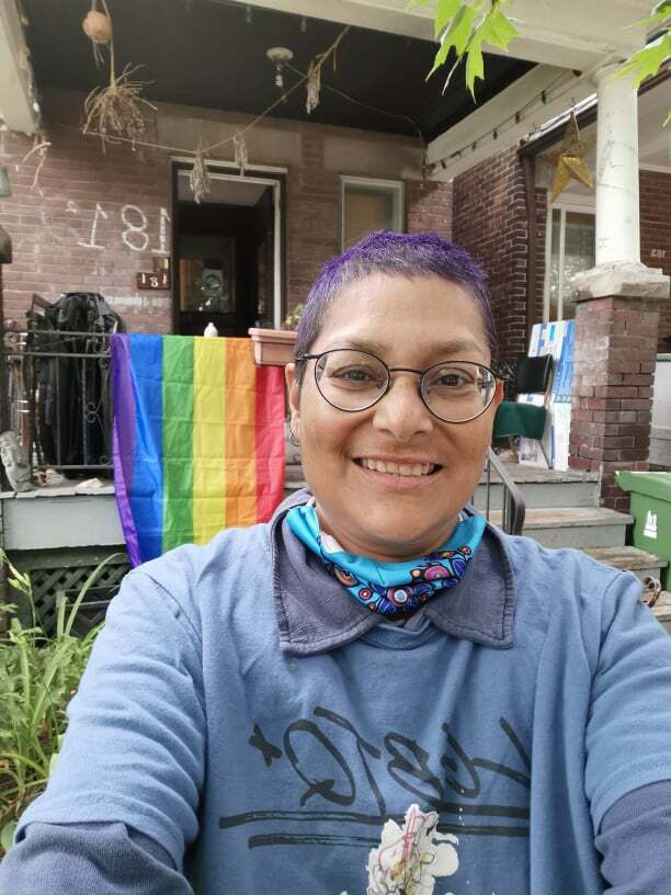 A smiling person wearing glasses in front of the porch of a brick building with a rainbow flag hung over the railing