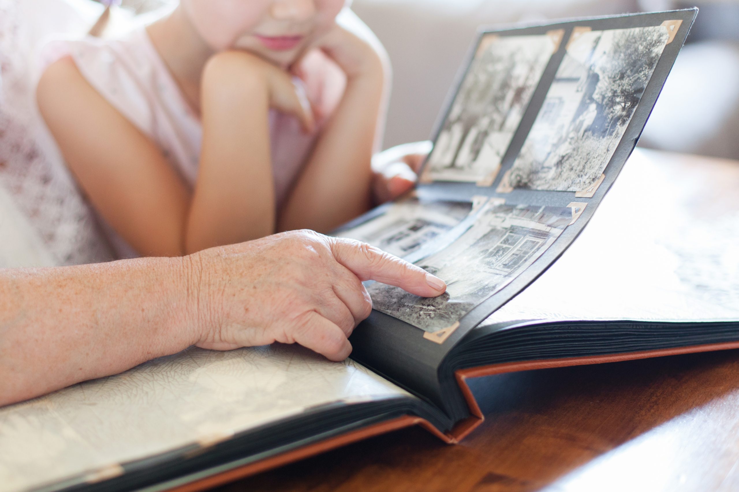 the hand on an elderly person pointing to a old black and white photo in a photo album resting on a table with a young person looking on