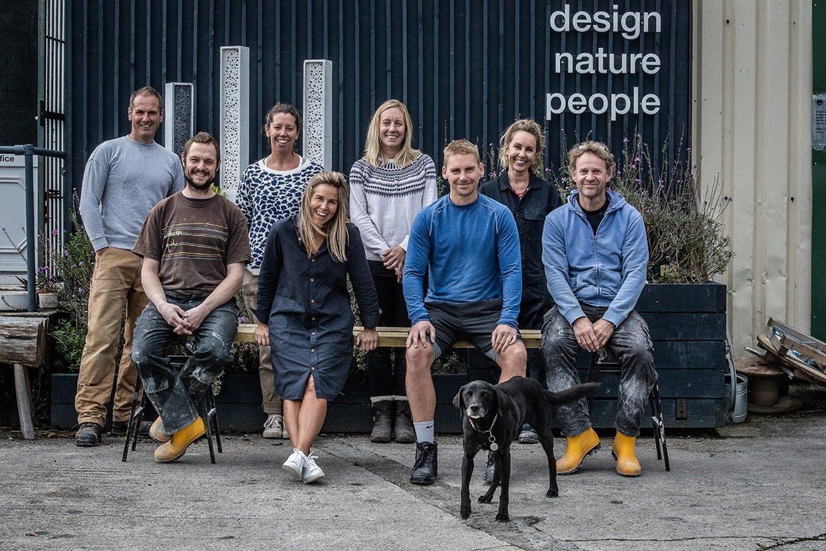 A group of eight women and men and a black dog from Green@Blue sitting and standing outside on front of a corrugated metal wall and three large bee bricks. The words design nature people appear on the photograph.