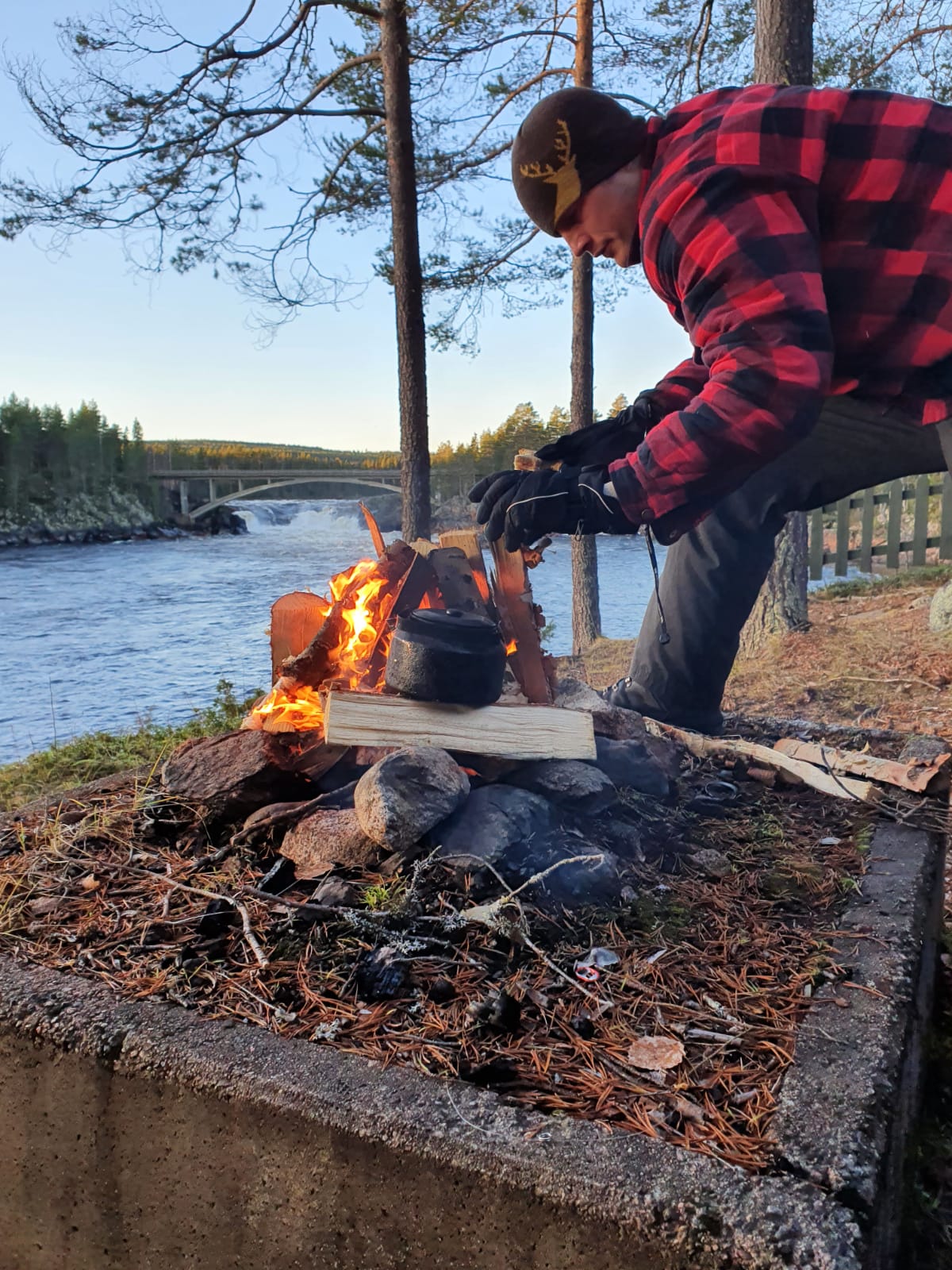 The Unplugged host Craig Craig Gavigan adds wood to a campfire in the woods by the side of a wide river with woodland and a bridge spanning small waterfalls in the backdrop
