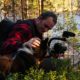 Podcaster Craig Gavigan of "The Unplugged Debate" sitting in the woods by a lake with a husky and holding a camera