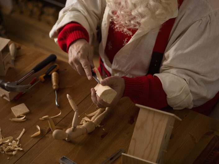 A pair of elderly hands hold a wooden toy and a carving tool. The hands belong to a person with a long white beard and dressed in red and white clothes. There are other wooden toys and carving tools on the wooden table on which the hands rest. Could this be Santa Claus, an elf or maybe even TranSanta ?
