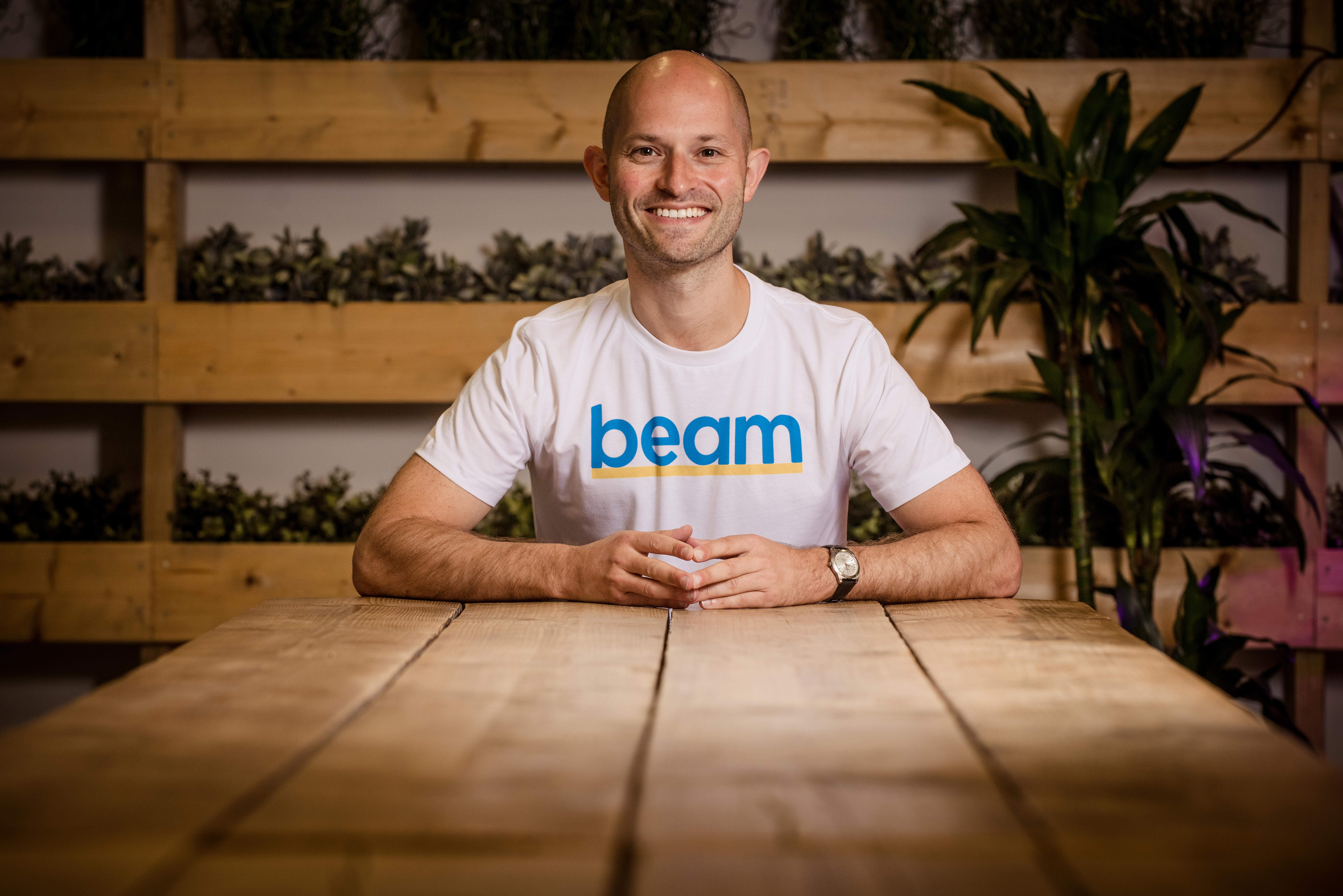 Alex Stephany, the founder of BEAM, the homeless crowdfunding platform, standing at what appears to be a wooden table directly facing the camera, with his arms and elbows resting on the wooden planks of the table, the lower part of his body is hidden from view by the table. He is smiling and wears a pale tee shirt with the word BEAM printed across his chest. In the background, out of focus, are wooden shelves filled with green plants and large green plants placed on the floor in back of him.