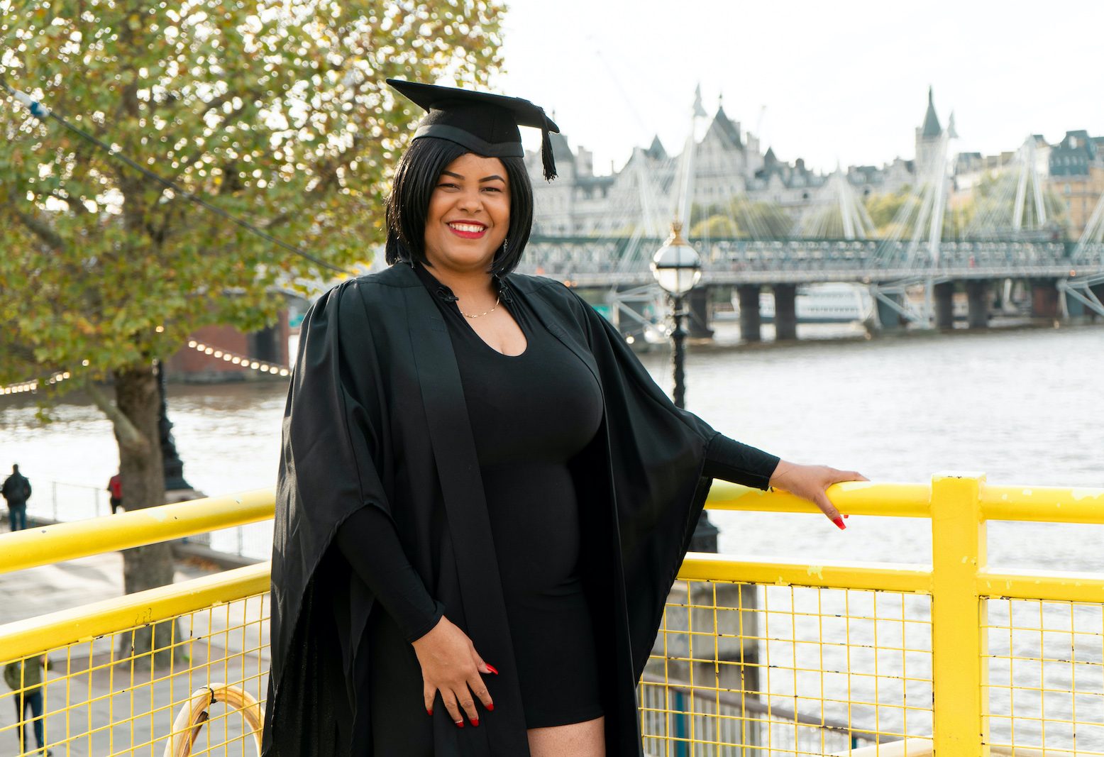 Smiling Beam member Decoda in a graduate's black cap and gown standing in front of London's Golden Jubilee Bridge and trees on the south bank of the Thames as she holds a yellow railing