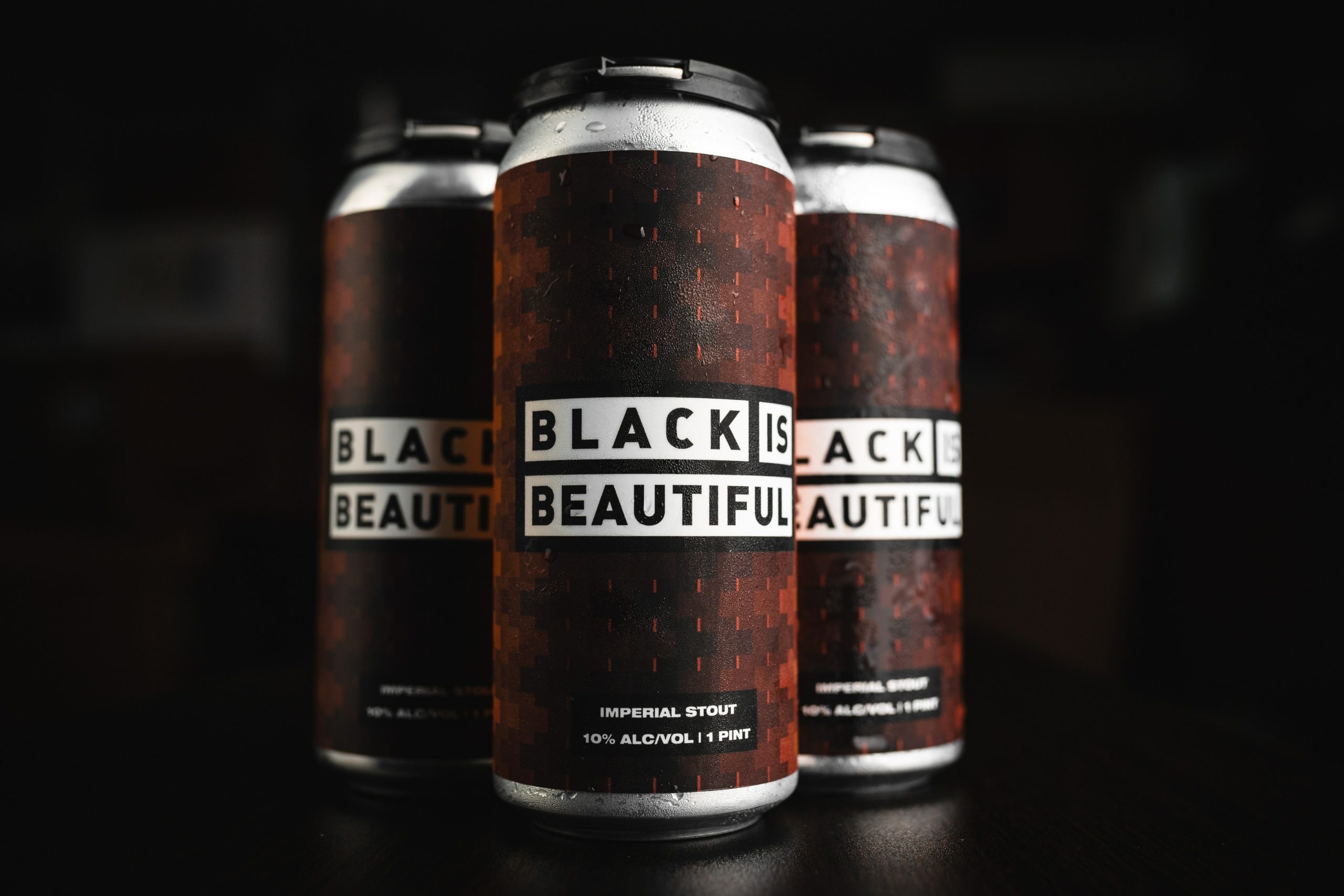 Three frosty aluminum beer cans on which are written the words "Black is Beautiful Imperial Stout 10% Alc/Vol". The cans are set against a black background.