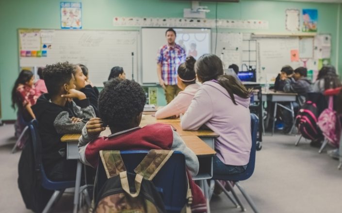 A classroom setting of adolescent students sitting at desks that are pushed together, facing a teacher who is standing in front of a variety of teaching materials - whiteboards, screens and posters - that are hanging on the wall