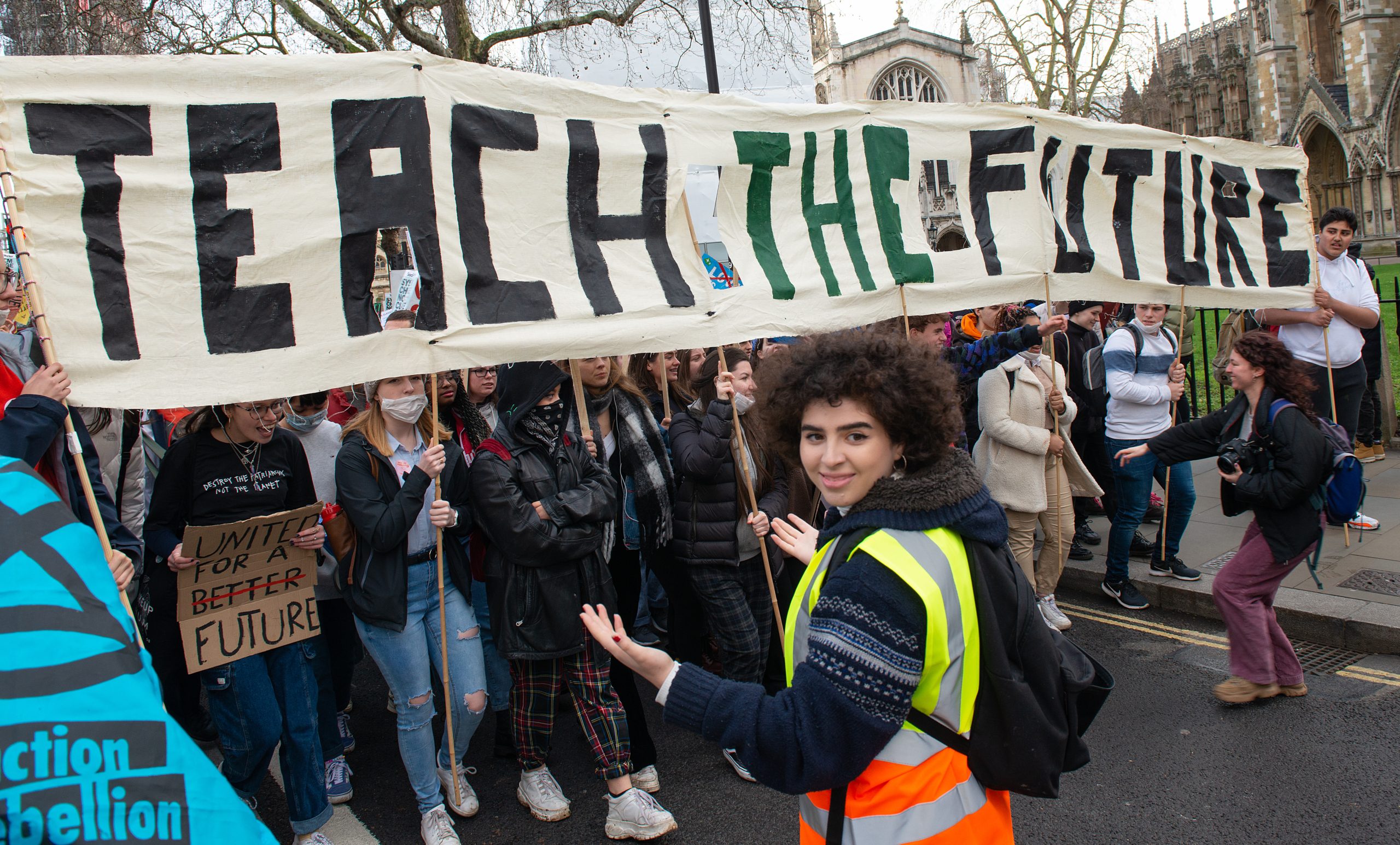Two youthful organisers, one wearing a reflective vest, stand in front of a wall of people standing in the street and on the sidewalk as they hold an unfurled banner on which is written "Teach The Future"