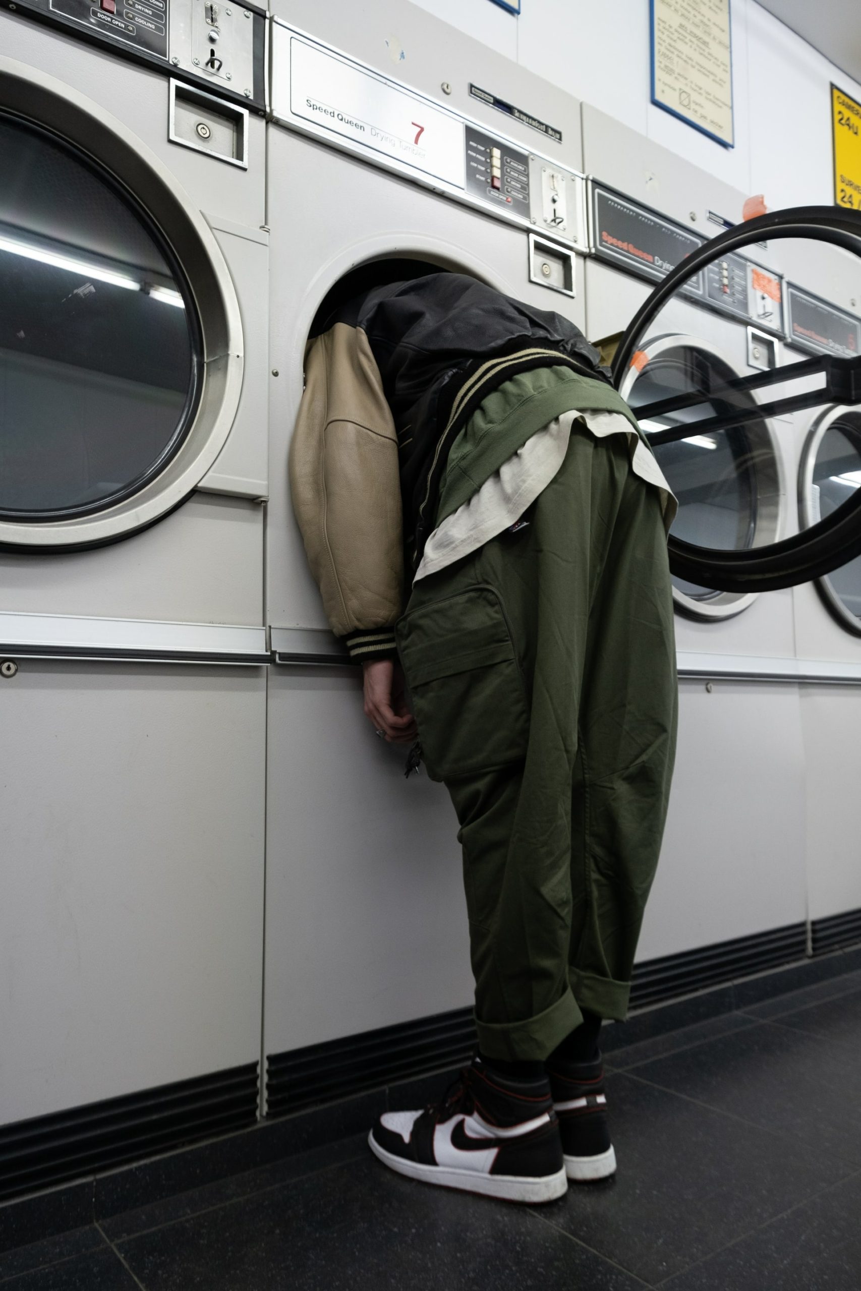 A person wearing green cargo pants, black and white running shoes and a old black vest with white sleeves, seen from behind as they place their head in a open washing machine in a laundrymat