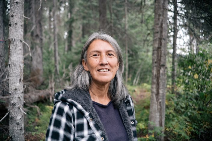 Wet’suwet’en Hereditary Chief Freda Huson wearing a plaid fleece jacket stands in front of a backdrop of forest trees