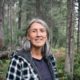 Wet’suwet’en Hereditary Chief Freda Huson wearing a plaid fleece jacket stands in front of a backdrop of forest trees