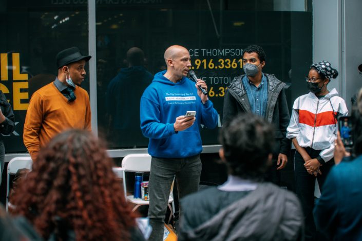 Four people including Phil Miller of FILSAA at CUNY Law school holding a mic and wearing a blue hoodie and jeans, standing in front of a crowd of people at an "Unlock the Bar" rally