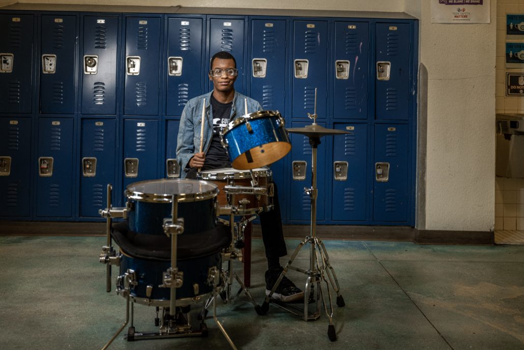 Student at guitars over guns playing the drums in front of lockers